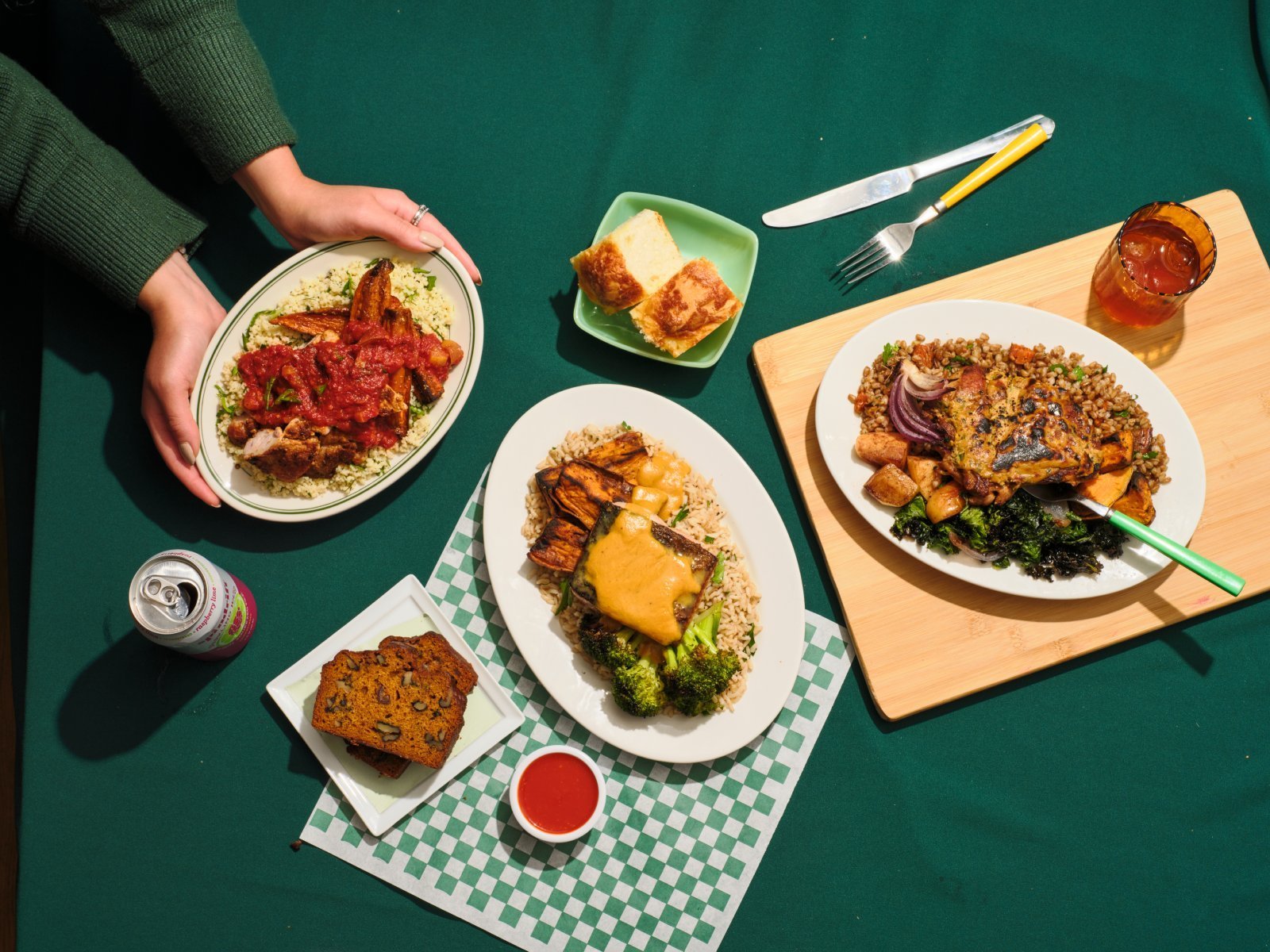 An overhead view of a fall dining table with several dishes. A person's arms, wearing a green sweater, are seen placing a plate of DIG’s chicken harissa on couscous with a spicy tomato sauce. Other dishes include roasted vegetables on rice, grilled chicken tray bake with kale, sweet potatoes, and lentils, as well as cornbread and a honey jar. A can of drink and various cutlery are also visible on the green checkered tablecloth.
